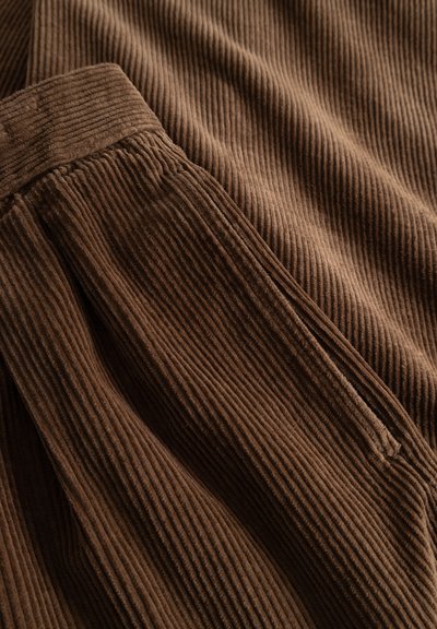 Brown corduroy fabric with visible vertical ridges and a close-up of a pocket with a waistband seam on textured trousers.