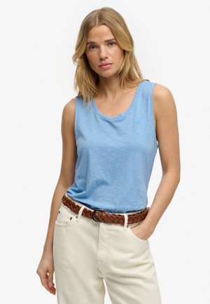 Femme aux cheveux blonds portant un haut sans manches bleu clair, un pantalon crème et une ceinture tressée marron, debout avec une main dans la poche.