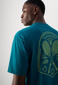 Teal bomulds T-shirt med grafisk tryk af krydsede tennisrackets og bolde i gul på ryggen. Kortærmet og design med rund hals.
