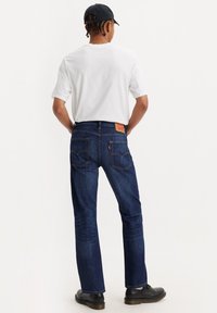 Vaqueros de mezclilla azul oscuro con costuras claras, de corte ajustado y dos bolsillos traseros. Combinados con una camiseta blanca simple y zapatos negros.