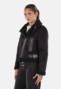 Giacca in shearling nera con colletto in pelliccia sintetica, tasche con cerniera e dettagli con fibbia. Presenta un design aderente e un tessuto testurizzato.