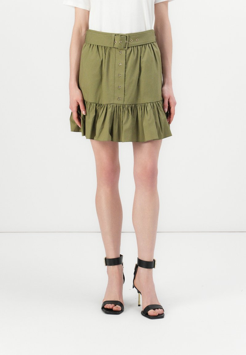 Gonna verde oliva con orlo arricciato, passanti per cintura e chiusura con bottoni frontali. Abbinata a sandali neri con tacco alto e cinturini alla caviglia.