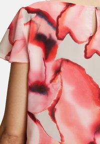 Blumenbluse mit transparenten, flatterhaften Ärmeln. Sie hat große pinke und rote abstrakte Blumenmuster auf einem hellen Hintergrund und ein Schlüsselaugen-Detail im Rücken.