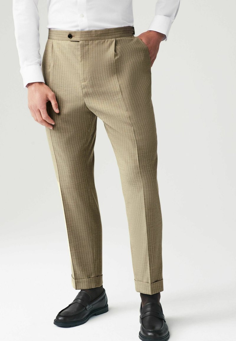 Pantaloni în dungi khaki cu un design frontal plat, textura materialului apare netedă, asortați cu mocasini negri, având o margine ușor tivită.