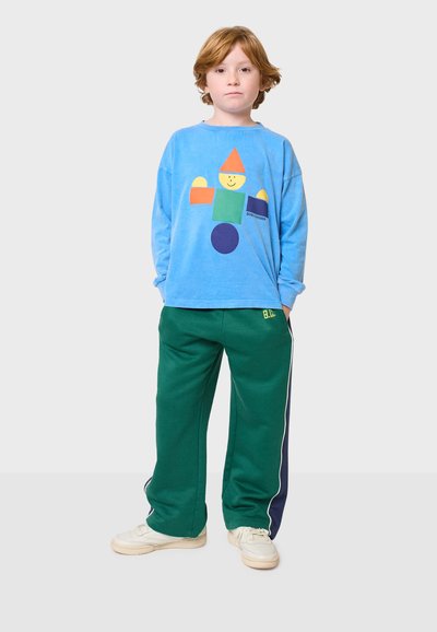 Svijetloplava majica dugih rukava s šarenim geometrijskim uzorkom, u kombinaciji sa zelenim trenirkama sa bočnim prugama i bijelim tenisicama.