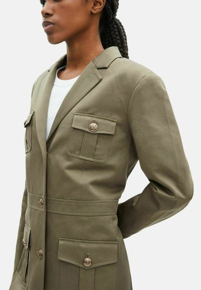 Frau mit geflochtenem Haar, die eine maßgeschneiderte olivgrüne Jacke mit goldenen Knöpfen und mehreren Fronttaschen über einem weißen Hemd trägt.