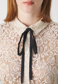 Gros plan d'une personne portant un chemisier en dentelle crème avec un motif floral et un petit ruban noir noué en nœud au niveau du col.