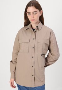 Beige pamut hosszú ujjú ing, két mellzsebbe, gombos elülső része van, fekete gombokkal; diszkrét fekete logó részlettel rendelkezik.