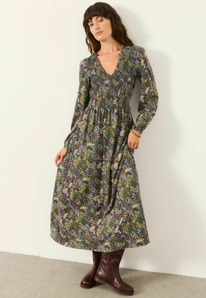 Frau trägt ein langes, geblümtes Kleid mit grünen, braunen und blauen Mustern, kombiniert mit dunkelbraunen Stiefeln bis zur Mitte der Wade, steht vor einer weißen Wand.