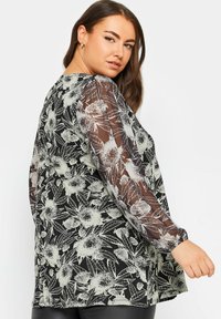 Blusa nera floreale con maniche trasparenti, caratterizzata da motivi floreali bianchi e una vestibilità rilassata. Tessuto testurizzato con polsini arricciati.