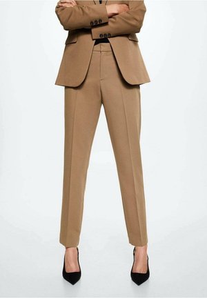 Persona vestida con un traje a medida color canela con pantalones cortos y tacones negros de punta, de pie con los brazos cruzados frente a un fondo blanco.