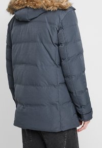 Chaqueta acolchada gris oscuro con capucha forrada de piel, que presenta costuras acolchadas horizontales y puños elásticos para proporcionar calidez y comodidad.