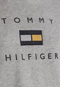 Szara bawełniana tkanina z czarnym haftowanym napisem "TOMMY HILFIGER" nad prostokątnym logo z czarnymi, szarymi i żółtymi paskami.