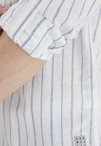 Camisa de lino blanca con rayas verticales azules, con puños enrollados y una pequeña etiqueta en la parte inferior derecha. Tejido ligero y texturizado.