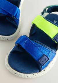 Blaue Kindersandalen mit grünen Riemen, strukturiertem Stoff, verstellbaren Schnallen und gesprenkelten weißen Gummisohlen.