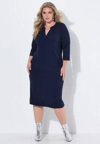 Navyblaues Kleid mit V-Ausschnitt, Dreiviertelärmeln, seitlichen Taschen und geradem Schnitt. Das Model trägt silberne Ankle Boots.