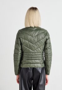 Olivenfarget pufferjakke med et chevron-quiltet design. Har høy krage, tettsittende silhuett og glinsende tekstur. Kombinert med svarte bukser.