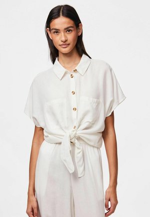 Haut blanc à boutons avec des manches courtes, noué à la taille. Il dispose de deux poches avant et de boutons en bois, fabriqué à partir d'un tissu léger.