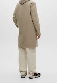 Abrigo de lana beige con capucha, corte recto y ventilación trasera. Combinado con pantalones cargo crema y zapatillas negras con acentos blancos.
