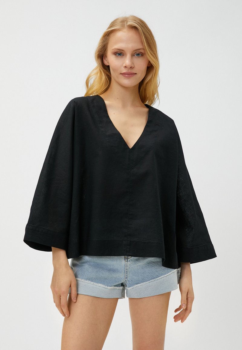 Schwarzes Leinenoberteil mit V-Ausschnitt und weiten Dreiviertelärmeln. Das Design ist locker geschnitten und wird mit hellblauen Jeansshorts kombiniert.