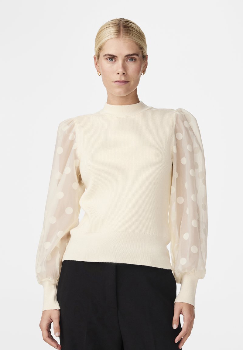 Pull en crème à côtes avec des manches bouffantes transparentes à pois et un col montant. L'ourlet du bas et les poignets sont ajustés et unis.