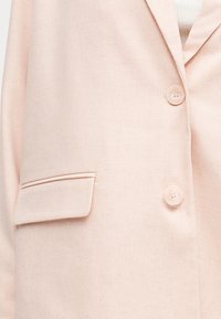 Απαλό ροζ σακάκι με δύο στρογγυλά κουμπιά και τσέπη με καπάκι στην αριστερή πλευρά, φορεμένο πάνω από λευκό ριμπ πλεκτό τοπ.