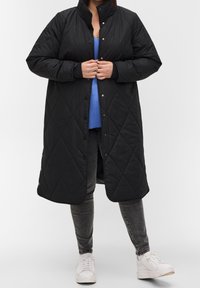 Sort lang quilted jakke med høj krave og trykknaplukninger, båret over en blå top og parret med grå jeans og hvide sneakers.