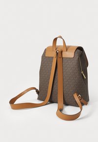 Sac à dos en toile brun monogrammé avec des accents en cuir beige, doté de sangles réglables, d'une poignée supérieure et d'une poche latérale zippée.