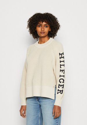 Maglione a maglia color crema con scollatura a costine e maniche lunghe. Presenta un logo "HILFIGER" blu sulla manica sinistra. Abbinato a jeans blu.