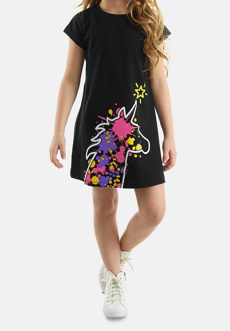 Fille portant une robe noire avec un motif licorne éclaboussé de peinture colorée et des baskets blanches, debout avec une jambe croisée.