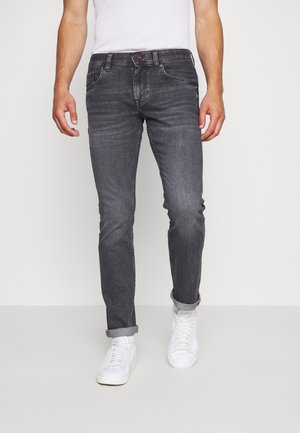 Mann trägt schlanke, dunkelgraue Jeans mit hochgekrempeltem Saum und weiße Sneaker, steht vor einem einfarbigen hellen Hintergrund.