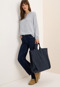 Sac en toile bleu marine avec doubles poignées, présentant des lettres en relief subtiles et un design robuste, associé à un chemisier à rayures légères et un jean foncé.