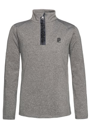 Jongen staat en draagt een grijze pullover met een kwartrits, groene waterdichte broek en zwarte laarzen tegen een effen lichte achtergrond.