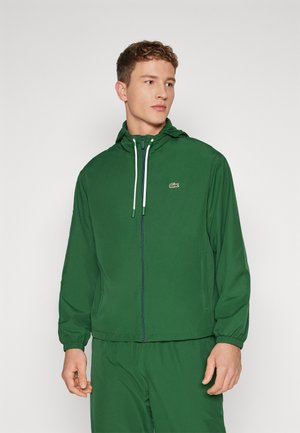 Chaqueta ligera verde con capucha, cremallera frontal y puños elásticos. Presenta un pequeño logo en el lado izquierdo del pecho, textura suave y diseño casual.