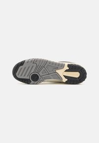 Semelle de chaussure de sport noire et beige avec caoutchouc texturé, présentant un motif géométrique et un détail de logo. Le design est structuré pour une adhérence optimale.