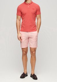 Homme portant un polo rouge ajusté, un short rose clair et des mocassins en cuir marron foncé, debout avec une main dans la poche.