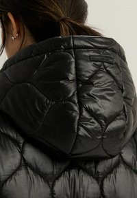 Czarna kurtka puchowa z pikowanym wzorem, błyszczącą fakturą i dopasowaną kapturem z naszywką logo na plecach.