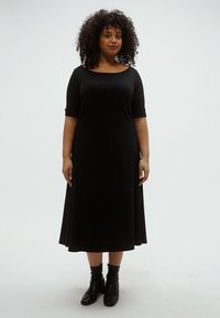 Rochie neagră, până la genunchi, cu un decolteu rotund și lejer și mâneci trei sferturi. Material moale și neted, cu o siluetă fluidă. Ghete negre până la gleznă.