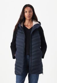 Marineblauwe gewatteerde gilet met capuchon, voorzien van verticale gestikte textuur, ritssluiting en beige voering. Draagt men over een zwarte gebreide trui.