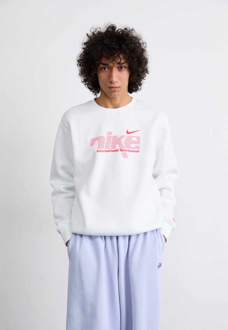 Sweatshirt branco com estampa gráfica rosa que diz "nike International Sportswear". Combinado com calças soltas em lavanda. Tecido suave, decote redondo.