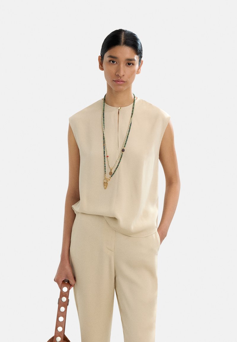 Beżowy top bez rękawów z przodem z guzikami, zestawiony z dopasowanymi spodniami. Modelka trzyma brązową torbę z okrągłymi wycięciami oraz naszyjnikami warstwowymi.