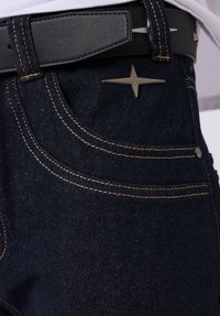 Jeans de mezclilla azul oscuro con costuras amarillas, un adorno de estrella metálica en el bolsillo delantero y un cinturón de cuero negro con hebilla plateada.