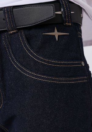 Jeans de mezclilla azul oscuro con costuras amarillas, un adorno de estrella metálica en el bolsillo delantero y un cinturón de cuero negro con hebilla plateada.