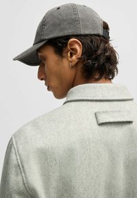 Gray wool coat with a folded collar and subtle texture, featuring a stitched "HUGO" label on the back. Complemented by a gray cap.