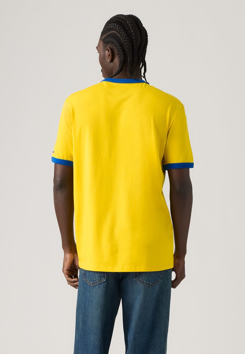 Homme aux cheveux tressés portant un t-shirt jaune avec un col et des bords de manches bleus, et un jean bleu, tourné de dos à la caméra.