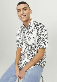 Chemise à manches courtes blanche avec motif floral noir, col boutonné et coupe décontractée, associée à un jean en denim bleu clair.