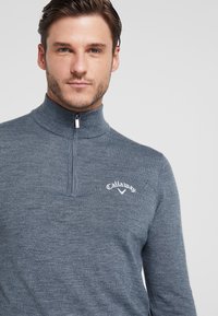 Suéter cinza com zíper no pescoço, feito de tecido tricotado. Apresenta um colarinho alto e o logótipo "Callaway" bordado no peito. Textura suave, design ajustado.