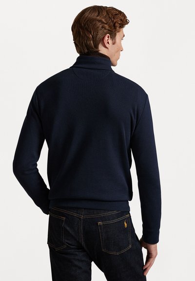 Marineblå pull med høy krage, laget av myk stoff. Kombinert med mørke denimjeans med synlig søm og et logo på baklommen.