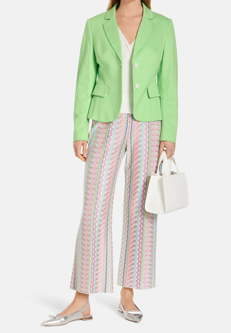 Blazer ajusté vert clair avec deux poches avant, porté sur un haut blanc ; associé à un pantalon large à motifs dans des couleurs pastel et des ballerines argentées.