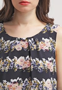 Donna che indossa un vestito nero senza maniche con motivi floreali orizzontali in tonalità di rosa, viola, bianco e verde.